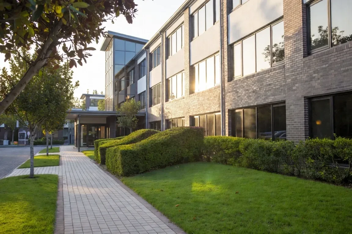 Modern brick and glass office building with a tree-lined path and manicured hedges in warm sunset light