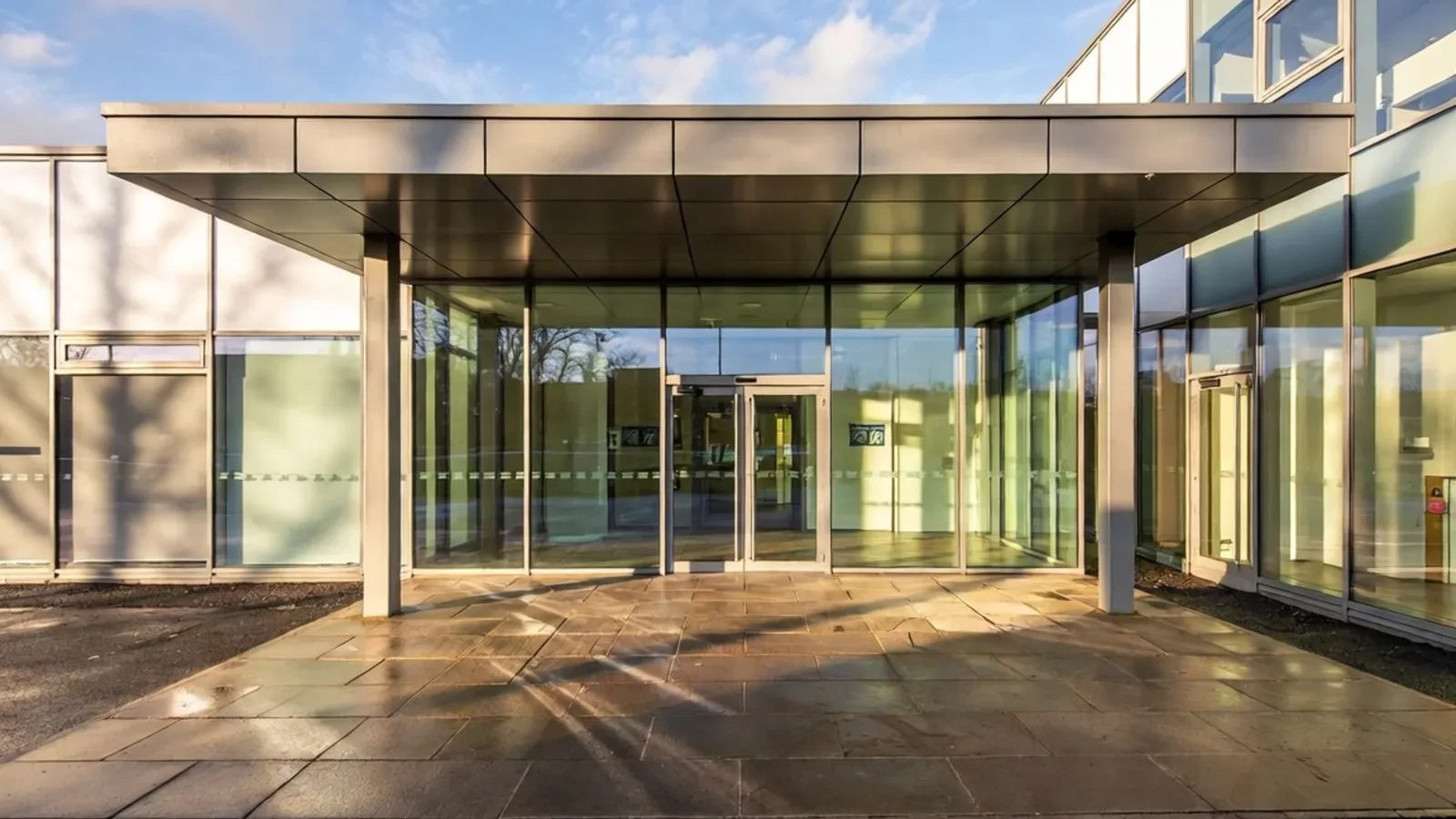 Modern office entrance with glass canopy and stone paving at dusk